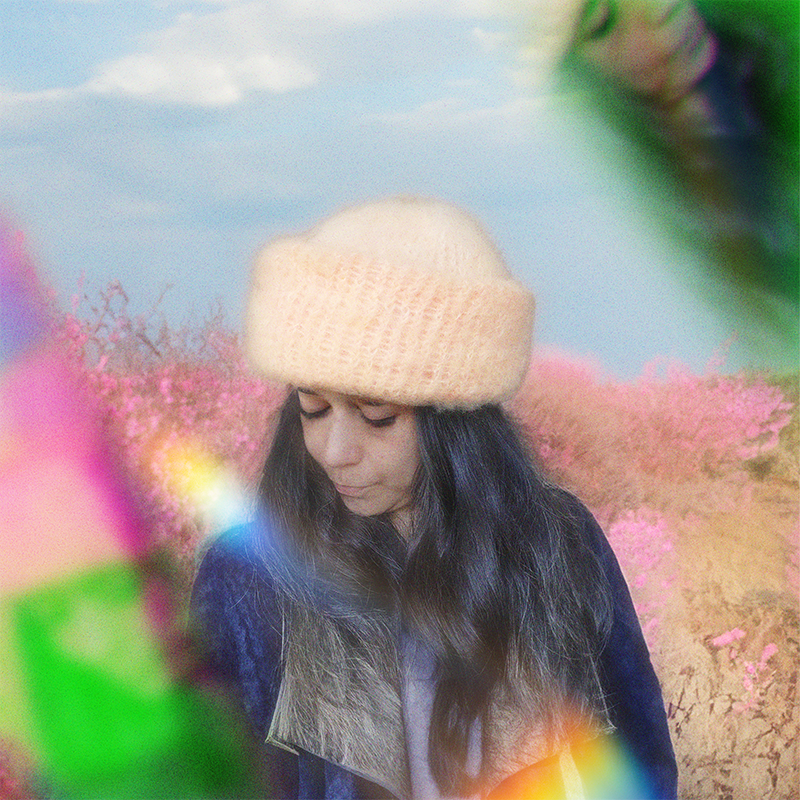 Person wearing a peach hand-knitted mohair beanie, with a pink and floral background, surrounded by a prism effect.