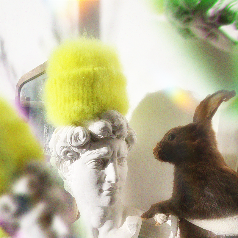 Yellow hand-knitted mohair beanie on a statue with a rabbit looking at it, surrounded by a prism effect.
